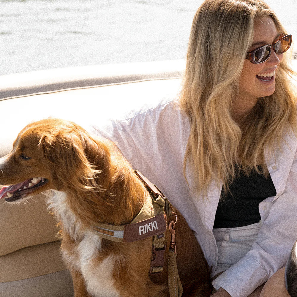 Woman smiling while sitting on a boat beside her brown and white dog wearing our personalised harness labeled “RIKKI”.