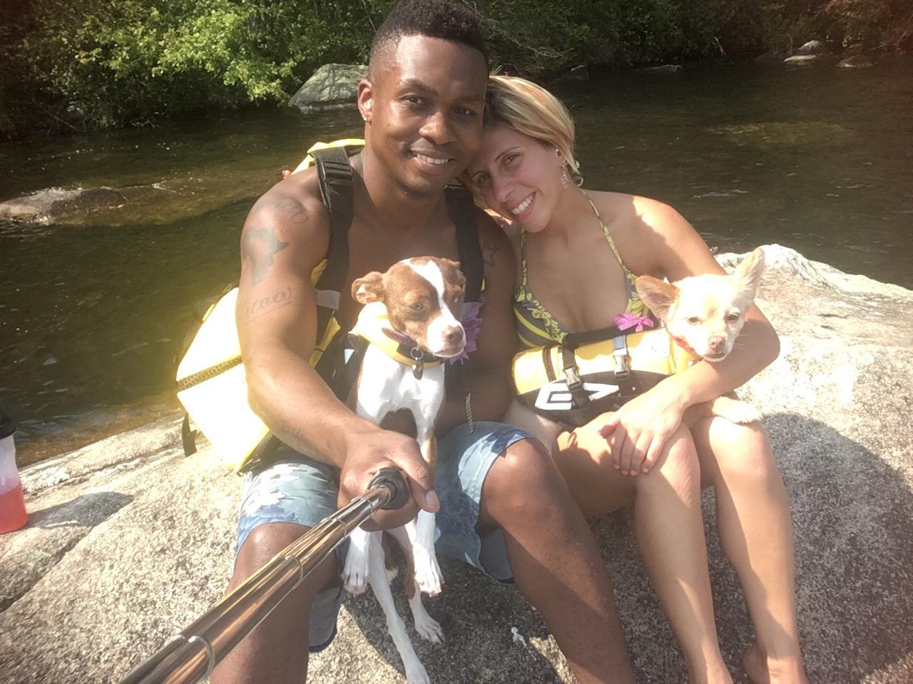 Two people sitting on a rock by a river with two small dogs.